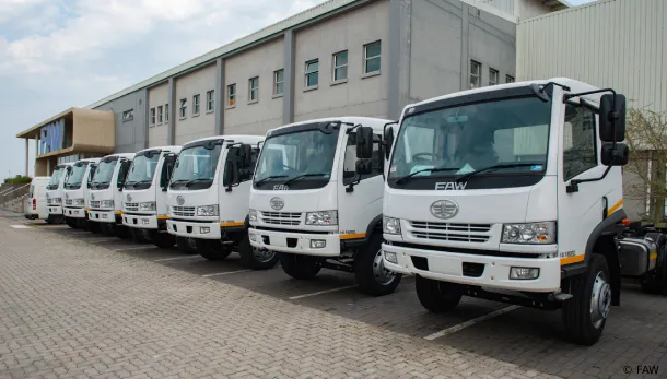 A fleet of new white FAW commercial trucks parked in a row outside a factory warehouse building.