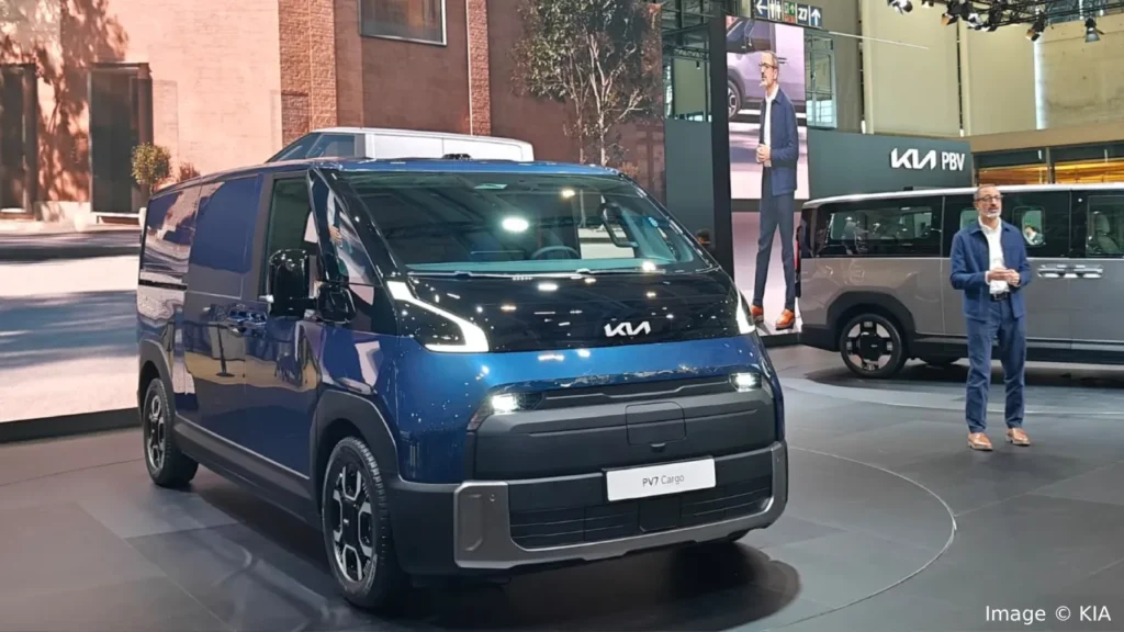 Deep blue Kia PV7 Cargo electric van displayed on a rotating stage at a Kia PBV motor show event with a presenter standing in the background.