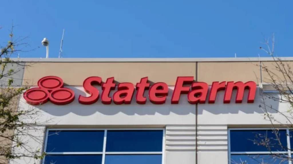 A red three-dimensional State Farm brand sign and three-oval logo emblem mounted on the beige exterior wall of a modern commercial office building above large glass windows against a clear blue sky.