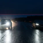 A photorealistic night road scene showing an oncoming vehicle's bright LED headlights causing intense highway glare alongside a secondary car demonstrating adaptive beam technology by casting a shaded cut-out area over the road surface to prevent blinding oncoming traffic.