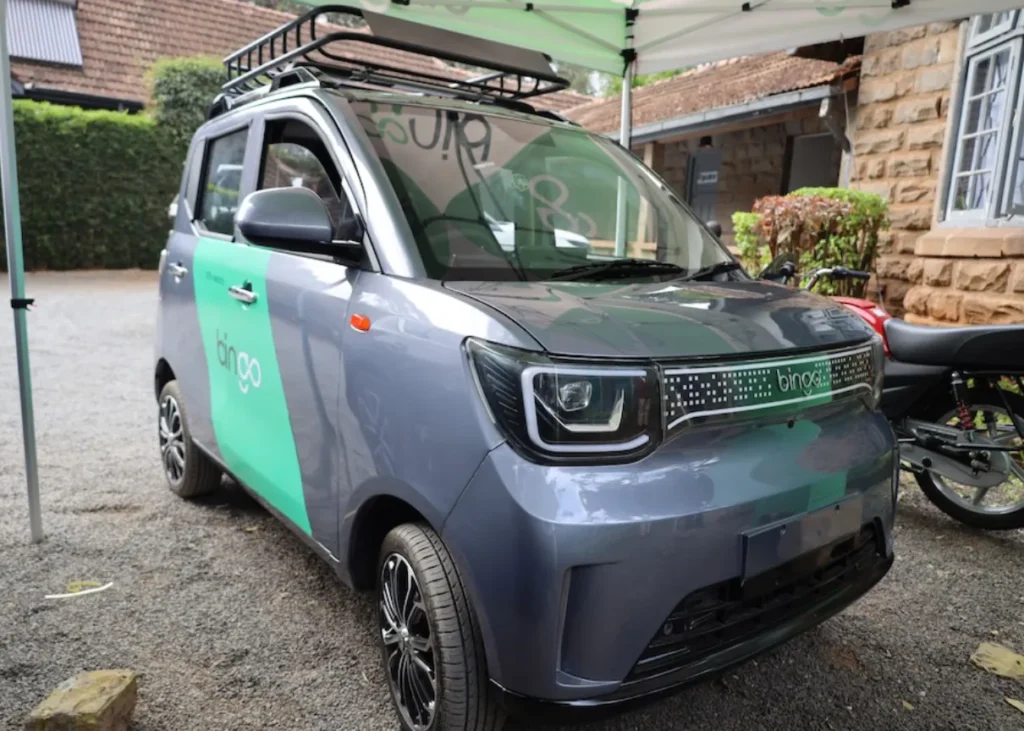Close-up front three-quarters view of a grey Bingo E2 electric microcar with a roof rack, parked outdoors under a canopy next to a motorcycle.