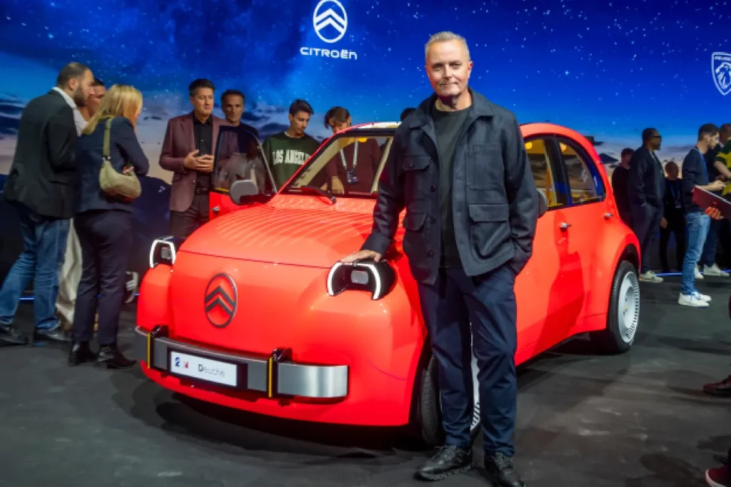 • A middle-aged male car designer dressed in a black jacket and dark trousers standing next to the bright red Citroën 2CV electric concept car at a busy auto expo showroom floor.