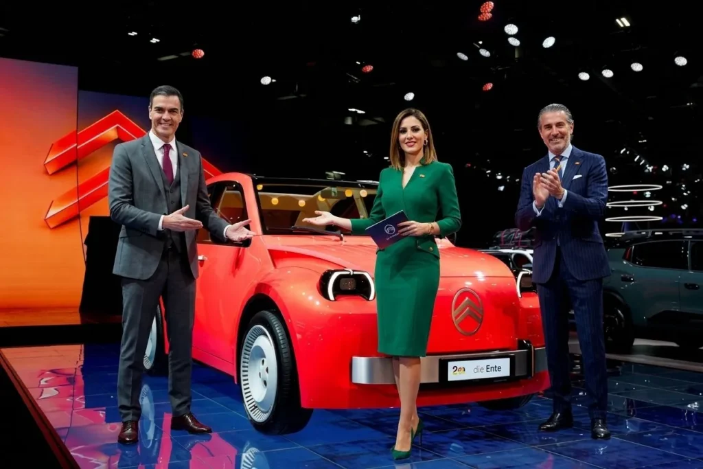 A bright red modern Citroën 2CV electric concept car being unveiled on a high-tech showroom stage by two sharply dressed Spanish male executives in business suits and one elegant Spanish woman presenter wearing a professional emerald green dress.