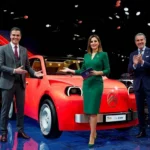 A bright red modern Citroën 2CV electric concept car being unveiled on a high-tech showroom stage by two sharply dressed Spanish male executives in business suits and one elegant Spanish woman presenter wearing a professional emerald green dress.