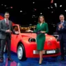 A bright red modern Citroën 2CV electric concept car being unveiled on a high-tech showroom stage by two sharply dressed Spanish male executives in business suits and one elegant Spanish woman presenter wearing a professional emerald green dress.
