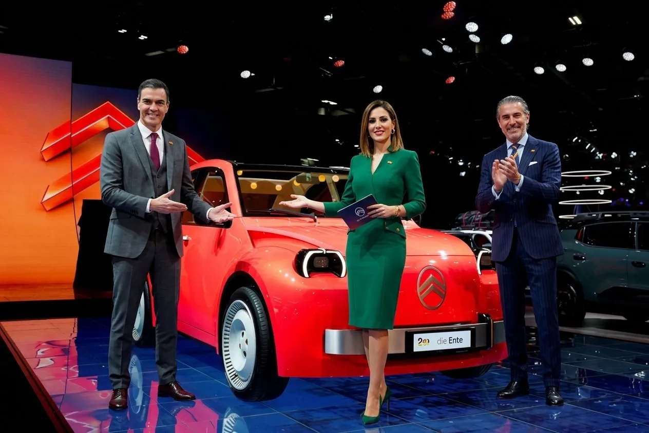 A bright red modern Citroën 2CV electric concept car being unveiled on a high-tech showroom stage by two sharply dressed Spanish male executives in business suits and one elegant Spanish woman presenter wearing a professional emerald green dress.