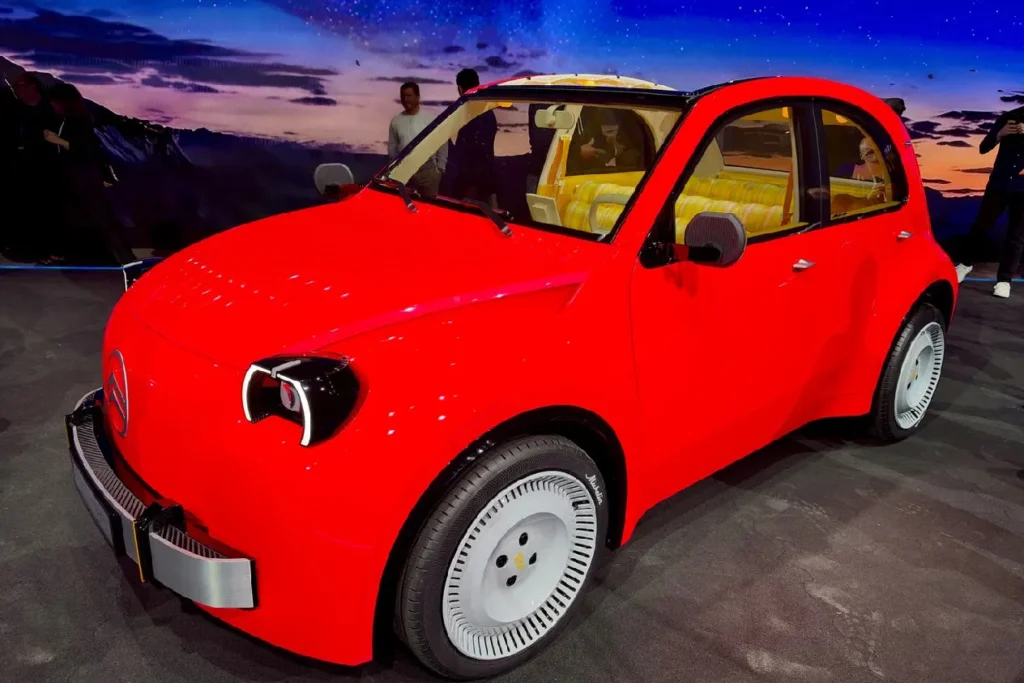A front three-quarter side-angle view of the bright red Citroën 2CV electric concept car, showcasing its unique white aerodynamic wheel rims, retro rounded cabin profile, and custom yellow striped interior seating.