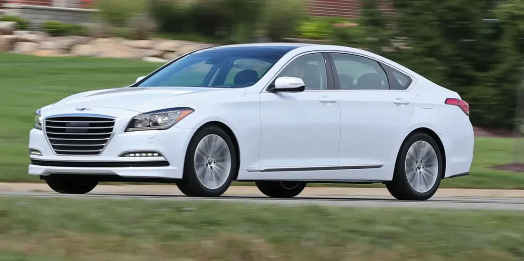 A side front three-quarter dynamic driving view of a pearl white 2017 to 2020 model Genesis G80 luxury sedan cruising on an asphalt road with motion-blurred green grass background.