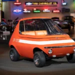 Side view of the bright orange 1969 GM XP-512E electric concept car displayed inside a showroom with classic car neon signs in the background.