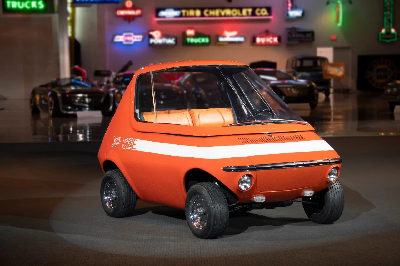 Side view of the bright orange 1969 GM XP-512E electric concept car displayed inside a showroom with classic car neon signs in the background.