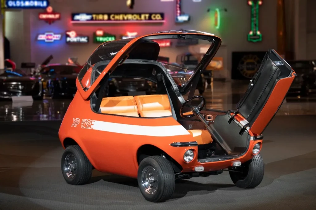 The orange 1969 GM XP-512E electric concept car with its front canopy and roof fully flipped open, revealing the two-seater cabin, orange vinyl seats, and steering wheel inside a museum showcase.
