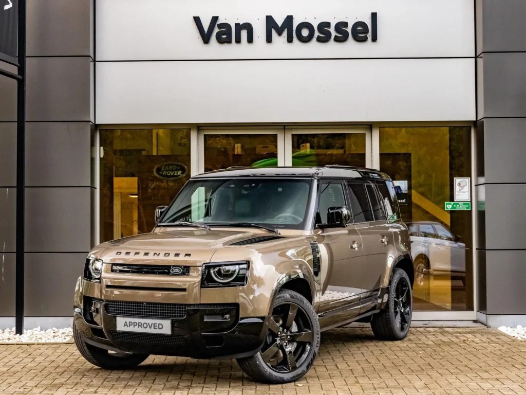 A front three-quarter dynamic shot of a luxury bronze and black Land Rover Defender 110 SUV parked outside a modern architectural building with dark textured walls.