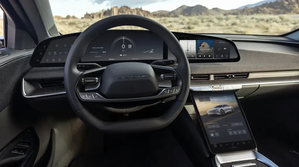 Close-up view of a Lucid Air dashboard featuring a steering wheel with the Lucid logo, a curved digital gauge cluster displaying zero miles per hour, and a swipeable audio and widget card interface on the right screen.