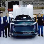 The dark green Nobo iNest 3.0 transforming electric concept car displayed at the IAA Mobility motor show booth in Germany, with automotive executives standing alongside the vehicle giving a thumbs-up gesture.