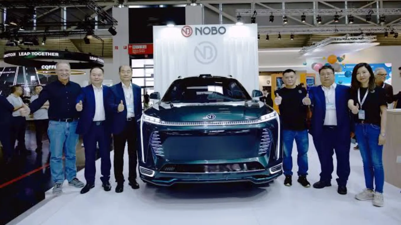 The dark green Nobo iNest 3.0 transforming electric concept car displayed at the IAA Mobility motor show booth in Germany, with automotive executives standing alongside the vehicle giving a thumbs-up gesture.