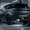 A Range Rover Sport SUV raised on a vehicle lift in a garage while a technician examines the rear aluminum subframe assembly for structural cracks related to NHTSA safety recall twenty-six-V-six-thirteen.