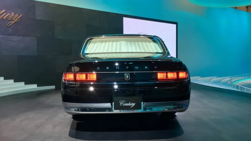 A Back profile view of a majestic glossy black Toyota Century ultra-luxury flagship limousine parked against a dramatic cloudy sunset sky, showcasing its executive wheelbase, signature multi-spoke chrome wheels, and phoenix emblems on the center caps.