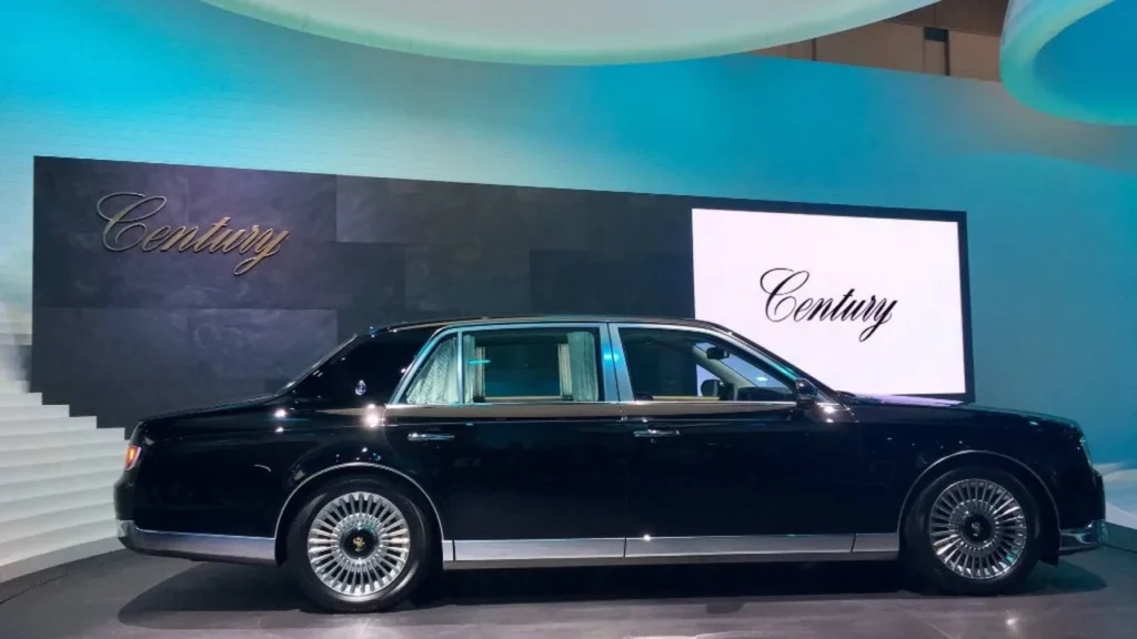 A long side profile view of a majestic glossy black Toyota Century ultra-luxury flagship limousine parked against a dramatic cloudy sunset sky, showcasing its executive wheelbase, signature multi-spoke chrome wheels, and phoenix emblems on the center caps.
