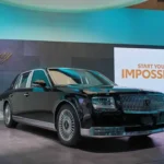A front view of a majestic glossy black Toyota Century ultra-luxury flagship limousine parked against a dramatic cloudy sunset sky, showcasing its executive wheelbase, signature multi-spoke chrome wheels, and phoenix emblems on the center caps.