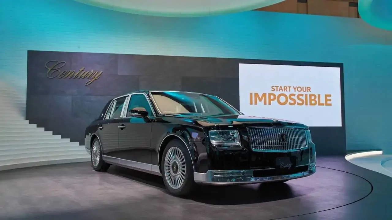 A front view of a majestic glossy black Toyota Century ultra-luxury flagship limousine parked against a dramatic cloudy sunset sky, showcasing its executive wheelbase, signature multi-spoke chrome wheels, and phoenix emblems on the center caps.