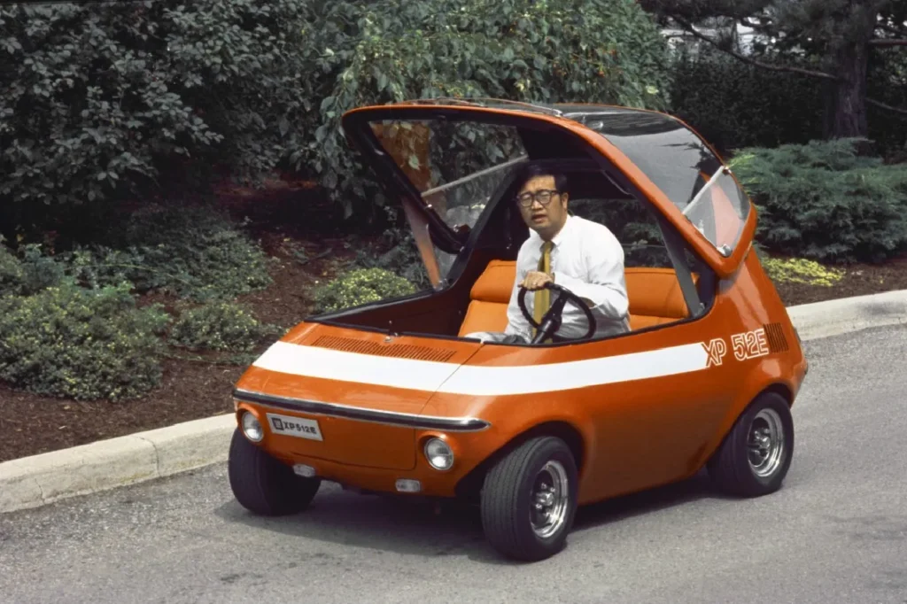 A vintage photograph of a man in a white dress shirt and tie sitting behind the steering wheel of the orange 1969 GM XP-512E electric concept car on an asphalt road with its glass canopy flipped open.