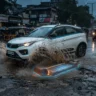 A white electric SUV driving through a deep pothole on a flooded Indian road during a monsoon dusk, showing a glowing cutaway diagram of the vulnerable flat battery pack under the floorpan.