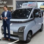 A light blue Wuling Air ev Lite micro electric car displayed at an international auto show booth, with two corporate professionals—one Chinese executive in a charcoal grey suit and one American executive in a navy blue suit—standing next to the vehicle discussing its specifications.