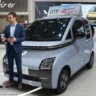 A light blue Wuling Air ev Lite micro electric car displayed at an international auto show booth, with two corporate professionals—one Chinese executive in a charcoal grey suit and one American executive in a navy blue suit—standing next to the vehicle discussing its specifications.