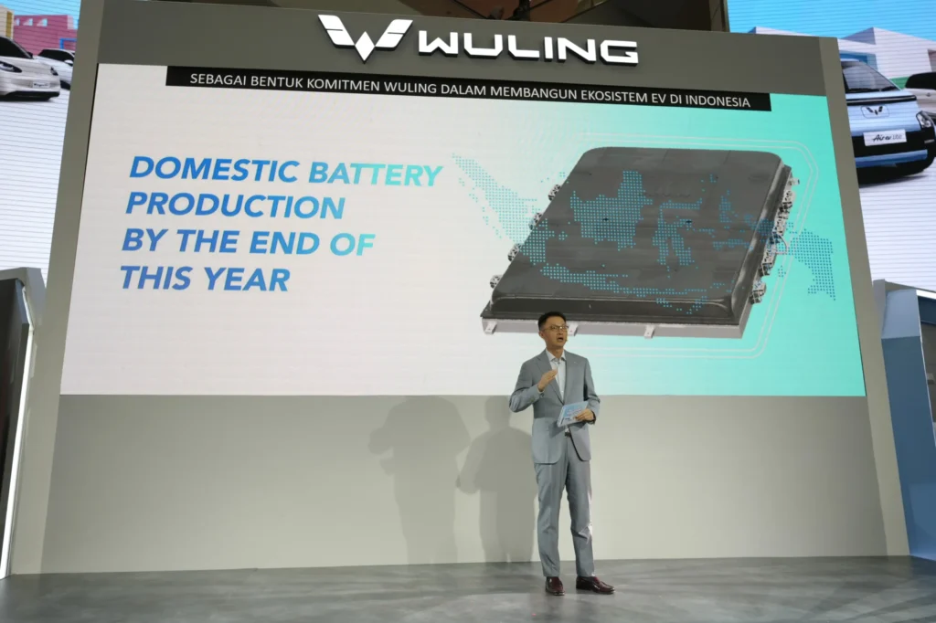 An executive in a light gray suit presenting on a motor show stage in front of a large Wuling presentation screen displaying an electric vehicle battery pack graphic with the text Domestic Battery Production By The End Of This Year.