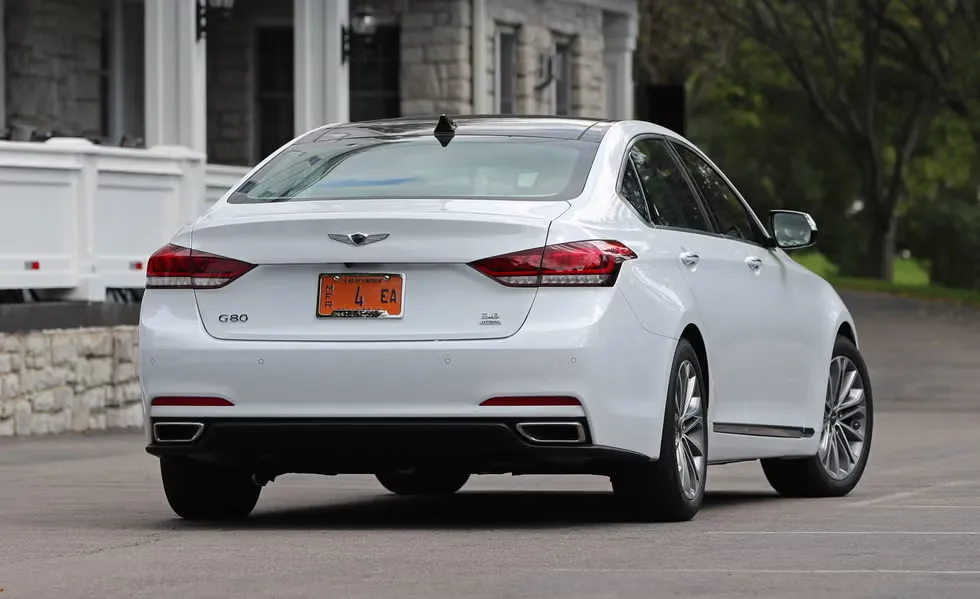 A rear three-quarter view of a pearl white 2017 to 2020 Genesis G80 3.8 AWD sedan parked outside a stone estate building, showcasing the dual exhaust tips, G80 and HTRAC badging, chrome rear wing emblem, and premium multi-spoke alloy wheels.